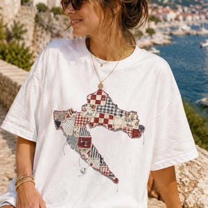 May include: A white short-sleeved t-shirt featuring a large, central graphic shaped like the map of Croatia. The map design is composed of various patchwork fabric squares in red, white, and blue patterns, including checks and floral prints.