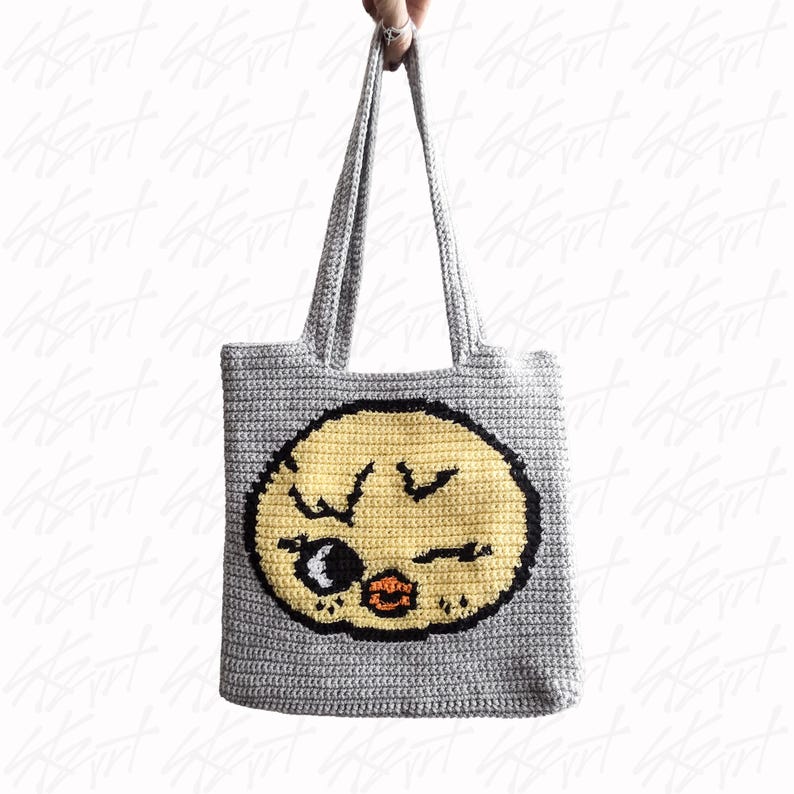 Puede incluir: Bolso tote de crochet gris con una gran cara de dibujos animados amarilla con contornos negros. La cara tiene un ojo gui&ntilde;ando y una boca fruncida. El bolso tiene dos asas grises.