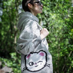 Puede incluir: Un bolso tote de ganchillo gris con la cara de un gato de dibujos animados, orejas rosas, bigotes negros y una expresi&oacute;n sonriente. El bolso se lleva al hombro y est&aacute; hecho de un material tejido texturizado. El fondo es una vista borrosa de follaje verde.