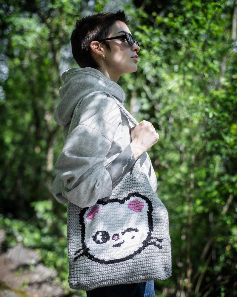 Puede incluir: Un bolso tote de ganchillo gris con la cara de un gato de dibujos animados, orejas rosas, bigotes negros y una expresi&oacute;n sonriente. El bolso se lleva al hombro y est&aacute; hecho de un material tejido texturizado. El fondo es una vista borrosa de follaje verde.