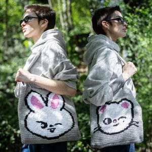Puede incluir: Dos bolsas tote grises con caras de animales de dibujos animados. Una bolsa presenta una cara de conejo blanco con orejas rosas, y la otra tiene una cara de gato blanco con orejas rosas. Ambas bolsas tienen contornos y correas negras, y son llevadas por personas con sudaderas grises.