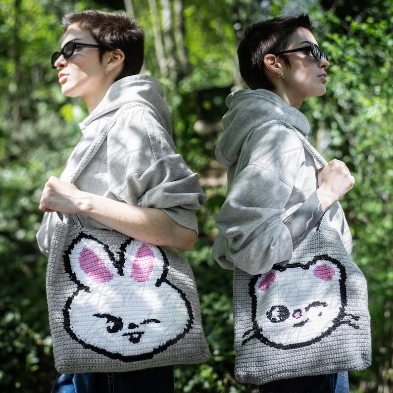 Puede incluir: Dos bolsas tote grises con caras de animales de dibujos animados. Una bolsa presenta una cara de conejo blanco con orejas rosas, y la otra tiene una cara de gato blanco con orejas rosas. Ambas bolsas tienen contornos y correas negras, y son llevadas por personas con sudaderas grises.