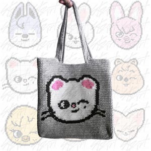 Puede incluir: Un bolso tote de crochet gris con una cara de oso de dibujos animados, orejas rosas y un contorno negro. El bolso tiene asas largas. El fondo presenta caras de dibujos animados pixeladas. El bolso est&aacute; sostenido por una mano.