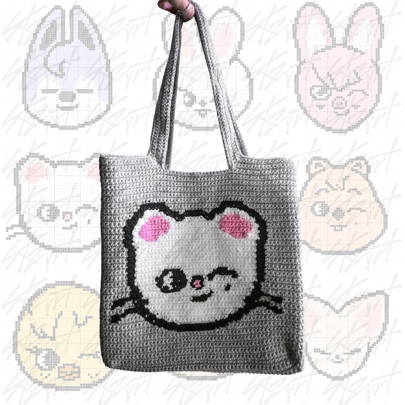 Puede incluir: Un bolso tote de crochet gris con una cara de oso de dibujos animados, orejas rosas y un contorno negro. El bolso tiene asas largas. El fondo presenta caras de dibujos animados pixeladas. El bolso est&aacute; sostenido por una mano.