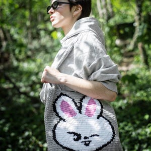 Puede incluir: Un bolso tote de crochet gris con una gran cara de conejo sonriente. El conejo tiene orejas rosas y contornos negros. El bolso se lleva al hombro, con una sudadera con capucha gris en el fondo. La cara del conejo es el punto focal del bolso.