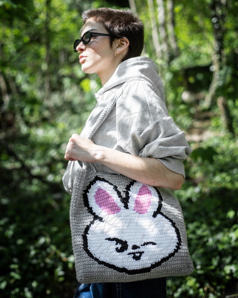 Puede incluir: Un bolso tote de crochet gris con una gran cara de conejo sonriente. El conejo tiene orejas rosas y contornos negros. El bolso se lleva al hombro, con una sudadera con capucha gris en el fondo. La cara del conejo es el punto focal del bolso.