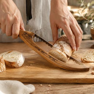 May include: A wooden bread slicer with a serrated metal blade, featuring an engraved chef hat icon and the text "Bake with love" on the handle. The tool is designed to guide the cutting of a crusty loaf of bread into even slices on a board.