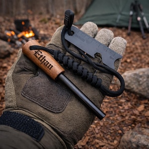 May include: A fire starter kit with a wooden handle engraved with the name "Ethan". The kit includes a black ferro rod, a black multi-tool with a ruler, and a black paracord lanyard. The items are held in a gloved hand.