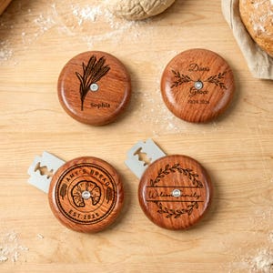 Könnte beinhalten: Vier Brotschneidewerkzeuge aus Holz mit eingravierten Designs. Eines zeigt Weizen, ein anderes einen Lorbeerkranz mit Namen und Datum, ein drittes mit einem Croissant und Text und das letzte mit einem Lorbeerkranz und Familiennamen. Jedes hat eine silberne Klinge.