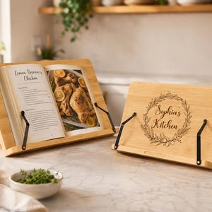 Peut inclure: Deux supports de livre de cuisine en bambou brun clair avec des porte-pages en métal noir. L'un soutient un livre de cuisine ouvert intitulé Lemon Rosemary Chicken, l'autre présente une gravure laser en couronne avec le texte Sophia's Kitchen.