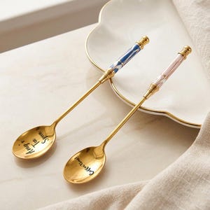 May include: Two gold-coloured spoons with decorative handles. One handle is blue and white, the other is pink and white. The spoon bowls have the phrases "Amy's Tea Spoon" and "Coffee time!" in black script. The spoons are on a white plate.