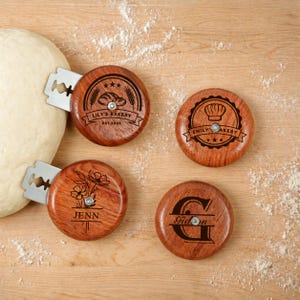 Könnte beinhalten: Vier Brotschneidewerkzeuge aus Holz mit eingebetteten Rasierklingen, jedes mit einem einzigartigen Design. Eines zeigt "Lily's Bakery" und "Est. 2023", ein anderes "Emily's Bakery", ein drittes mit dem Namen "Jenn" und das letzte mit dem Namen "Gideon".