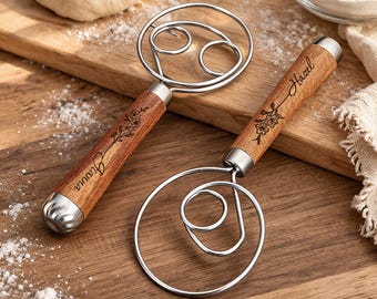 May include: Two stainless steel dough whisks with wooden handles. The handles are engraved with floral designs and names. One handle says "Gianna" and the other says "Hazel". The whisks are on a wooden cutting board.