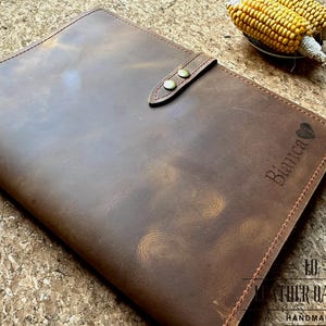 May include: A brown leather folio with a snap closure. The folio has a personalized engraving that reads "Bianca" with a heart symbol. The folio is handmade and has a distressed finish. The folio is a notebook or document holder.