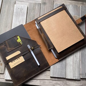 Puede incluir: Portafolio de cuero marrón abierto con múltiples compartimentos, incluyendo un bloc de notas, ranuras para tarjetas y un portalápices. El portafolio tiene un acabado envejecido y un interior beige. Un bolígrafo blanco está en el portalápices. La palabra "NOTES" es visible.