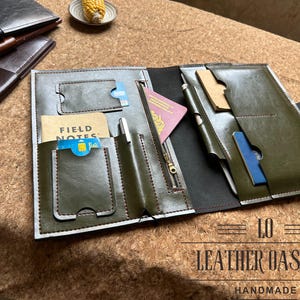 May include: Open olive green leather notebook with multiple pockets, a zipper, and a snap closure. The notebook contains a passport, cards, and a pen. The text "FIELD NOTES" is visible. The bottom of the image has the text "LEATHER OASISER HANDMADE".