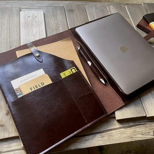 Puede incluir: Un portafolio de cuero marrón oscuro con un portátil, un bolígrafo y un bloc de notas. El portafolio tiene ranuras para tarjetas y un cierre de solapa. El portátil es plateado con un logotipo de Apple. El bloc de notas es negro.