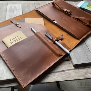 May include: Open brown leather portfolio with a pen, notepad, and a strap closure. The portfolio has multiple pockets and a pen holder. A small notepad with the words "FIELD NOTES" is visible.