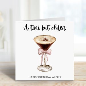 Puede incluir: Tarjeta de felicitación blanca con el texto "A tini bit older" sobre una ilustración de un martini. El martini tiene un lazo rosa y las palabras "HAPPY BIRTHAY ALEXIS" están en la parte inferior.