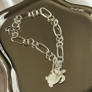 Puede incluir: Una pulsera plateada con un dije de tortuga. La pulsera presenta una cadena de eslabones ovalados y redondos alternos. El dije de tortuga es texturizado y detallado. La pulsera se exhibe en una bandeja de color plateado.