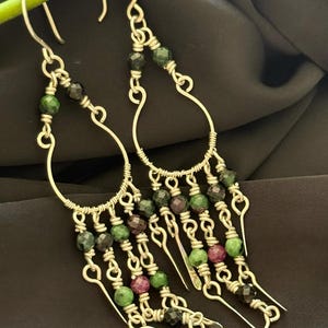Op de afbeelding: Zilverkleurige oorhangers met een druppelvormig ontwerp. De oorbellen hebben strengen groene, zwarte en kastanjebruine kralen. De kralen zijn geregen op zilverdraad, wat een gelaagde en gestructureerde uitstraling creëert.