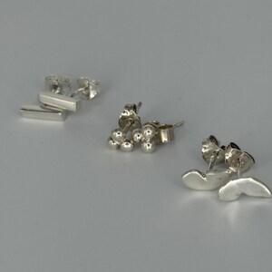 May include: Three pairs of silver stud earrings are shown against a light grey backdrop. The earrings include a pair of small bars, a pair of clustered spheres, and a pair of curved, wing-shaped designs. The earrings are all made of a shiny, metallic material.