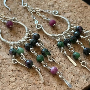May include: Silver chandelier earrings with dangling strands of faceted green, black, and pink gemstones. The earrings have a hook closure and are displayed on a cork surface.