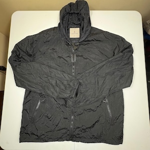 Puede incluir: Chaqueta negra con capucha, cremallera completa y dos bolsillos laterales. La chaqueta tiene una capucha con cordón y un pequeño parche con el logotipo en el pecho. El material parece ligero y ligeramente arrugado.