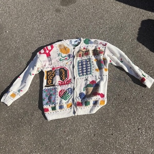 Könnte beinhalten: Eine cremefarbene Strickjacke mit langen Ärmeln, die mit verschiedenen bunten, strukturierten Aufnähern mit Näh- und Bastelmotiven wie Scheren, einer Nähmaschine, Garn und Herzen verziert ist und eine durchgehende Knopfleiste besitzt.