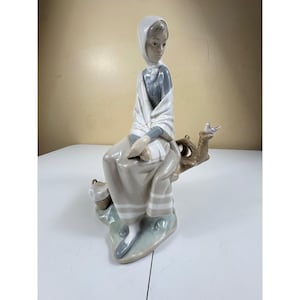 May include: A porcelain figurine of a seated woman in a white head covering and shawl, with a grey top and dress. She sits on a tree branch with a small bird. A basket is at her feet. The figurine is painted in muted tones of grey and white.