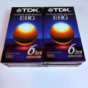Pode incluir: Duas cassetes de vídeo VHS TDK E-HG. A embalagem é azul escuro com um design de esfera laranja e amarela. O texto inclui "ULTIMATE PERFORMANCE", "EXTRA HIGH GRADE", "6 hrs" e "T-120".