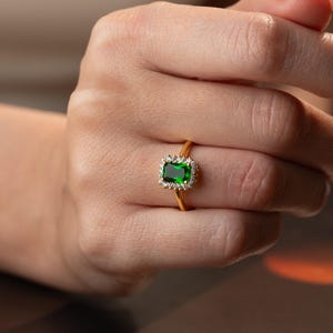 May include: A gold ring featuring a rectangular emerald-green center stone surrounded by a halo of small clear white stones. The band is thin and polished gold, showcasing a classic design with a vibrant green gemstone centerpiece.