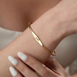 May include: A gold-coloured bangle bracelet with a small, elongated design in the centre. The bracelet has a smooth, polished finish and a simple, elegant design. The bracelet is worn on a wrist.
