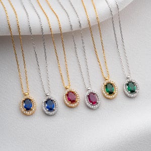 May include: A collection of gemstone pendant necklaces. The necklaces feature oval-cut gemstones in blue, red, and green, each surrounded by a halo of smaller stones. The chains are available in gold and silver tones, displayed on a white fabric.
