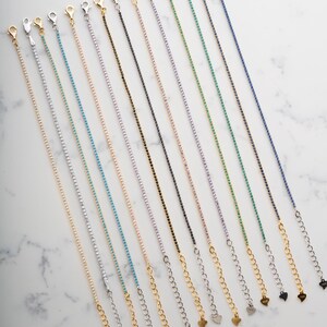 May include: A collection of delicate, colorful rhinestone bracelets with lobster claw clasps and heart-shaped charms. The bracelets are arranged on a white marble surface, showcasing a variety of colors including gold, silver, blue, green, and purple.