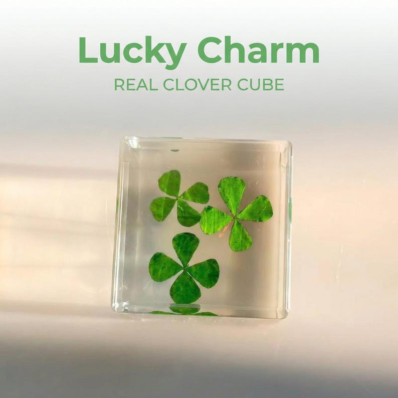 May include: A clear, square resin cube containing three green four-leaf clovers. The text "Lucky Charm" and "REAL CLOVER CUBE" are printed in green above the cube. The cube is displayed on a white surface.