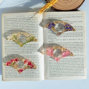 Puede incluir: Cuatro marcadores de resina con flores secas y láminas doradas se muestran en un libro abierto. Los marcadores tienen forma de abanico, con colores púrpura, verde, rosa y dorado. El libro está abierto en una página de texto.