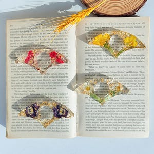 May include: Four clear resin butterfly-shaped bookmarks with dried flowers inside, resting on an open book. The bookmarks feature red, yellow, white, and purple flowers. A sprig of wheat is placed on top of the book.