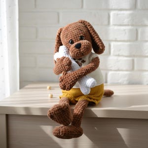 May include: A brown crocheted dog plush toy with floppy ears, wearing a cream-coloured shirt and yellow shorts. The dog is holding a white bone. The toy is sitting on a light-coloured surface.