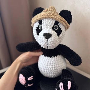 Może przedstawiać: Ręcznie robiony szydełkowy miś panda z czarno-białym futrem, w brązowym kapeluszu. Panda ma czarne łapy z różowymi podkładkami. Miś siedzi z wyciągniętymi ramionami i nogami.