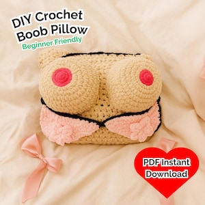 Puede incluir: Una almohada de crochet beige con pezones rosas y un diseño rosa debajo del busto. La almohada tiene ribetes negros y el texto "DIY Crochet Boob Pillow Beginner Friendly" es visible. También es visible un corazón rojo con el texto "PDF Instant Download".