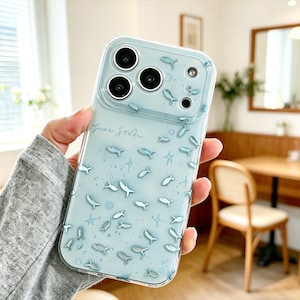 Puede incluir: Funda transparente para teléfono con fondo azul claro y un patrón de pequeños peces blancos y azules. La funda tiene un recorte para la cámara y la inscripción "Free Fish" en cursiva.