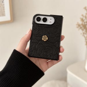 Könnte beinhalten: Eine schwarze Handyhülle mit strukturiertem Blumenmuster und einem kleinen goldfarbenen Rosenemblem in der Mitte. Die Hülle hat eine rechteckige Aussparung an der Oberseite für die Kameralinsen und den Blitz, passend für ein faltbares Gerät.