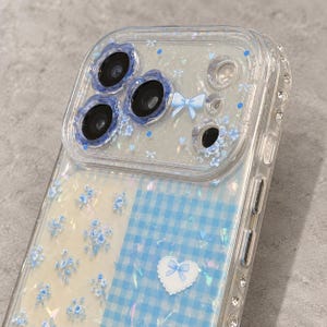 Könnte beinhalten: Eine transparente Handyhülle mit geteiltem Design, das auf einer Seite blaue Blumenmuster und auf der anderen ein blaues Vichy-Karo-Muster zeigt. Die Hülle enthält einen herzförmigen Aufkleber mit blauer Schleife sowie blumenförmige Linsenschutzringe.
