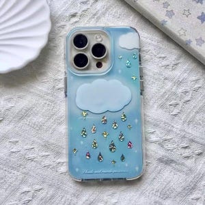 May include: A clear phone case with a blue sky design featuring white clouds, sparkling raindrops, and stars. The case has a transparent border and the text "Clouds and raindrops..." at the bottom.
