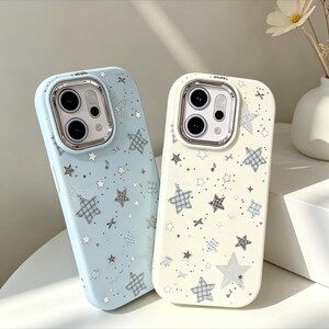 Puede incluir: Dos fundas para teléfono con un patrón de estrellas. Una es azul claro, la otra blanquecina. Ambas tienen un anillo plateado para la cámara y un patrón repetido de estrellas de varios tamaños y estilos.