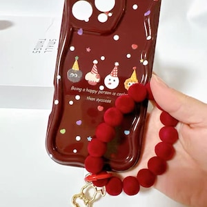 May include: A maroon phone case with a whimsical design featuring cartoon characters wearing party hats and the text "Being a happy person is cooler than success." A red beaded wrist strap with a gold-colored charm is attached.