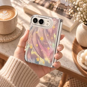 K&ouml;nnte beinhalten: Eine transparente, faltbare Smartphone-H&uuml;lle mit einem wirbelnden abstrakten Muster in Rosa-, Lila- und Goldt&ouml;nen, akzentuiert durch kleine gelbe Sterne und den Schriftzug "Star light" in zarter wei&szlig;er Schrift auf der R&uuml;ckseite.