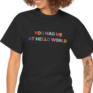 May include: Black t-shirt featuring the phrase "YOU HAD ME AT HELLO WORLD" in rainbow lettering. The top is a standard crew neck design.
