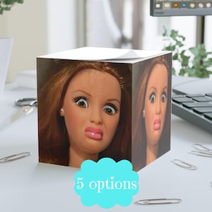 May include: A cube-shaped notepad featuring a doll's face with a surprised expression on multiple sides. The notepad has a light blue cloud-shaped graphic with the text "5 options". The background is a white desk.